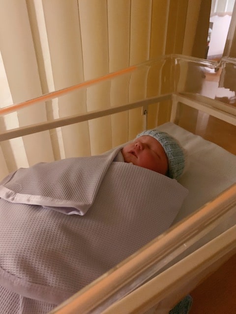 A newborn baby in hospital wrapped in a purple swaddle with a blue knitted hat